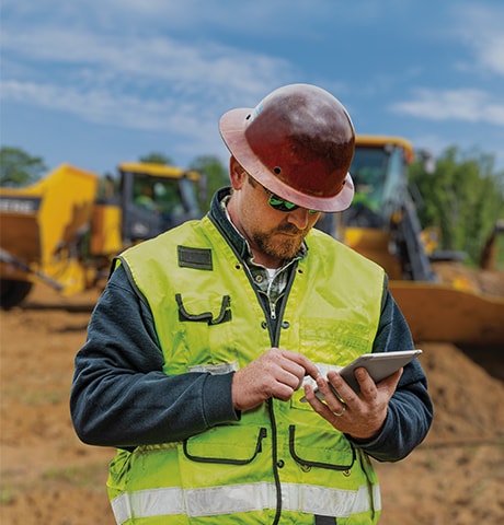 Ouvrier en construction portant une veste de sécurité verte et consultant son appareil mobile devant un chargeur et un tombereau.