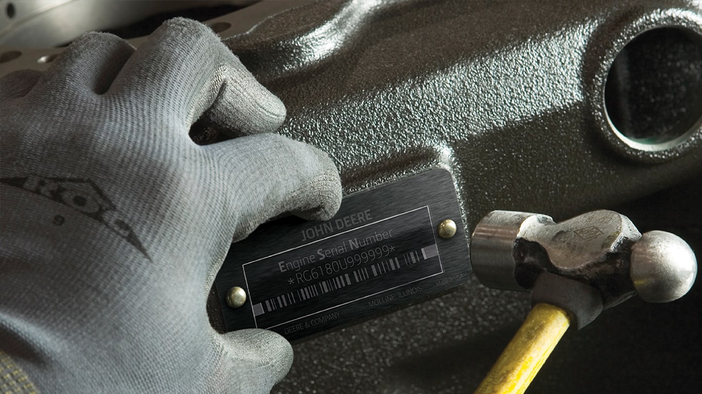 Personne portant un gant gris et tenant un marteau lors de la pose d’une plaque de numéro de série John Deere sur un moteur