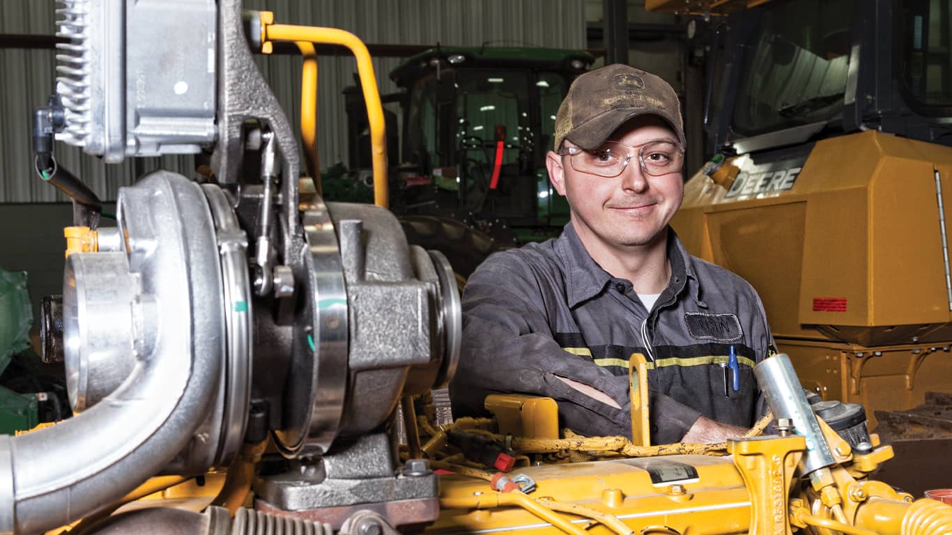 Homme portant des lunettes de sécurité et souriant à côté d’un moteur John Deere