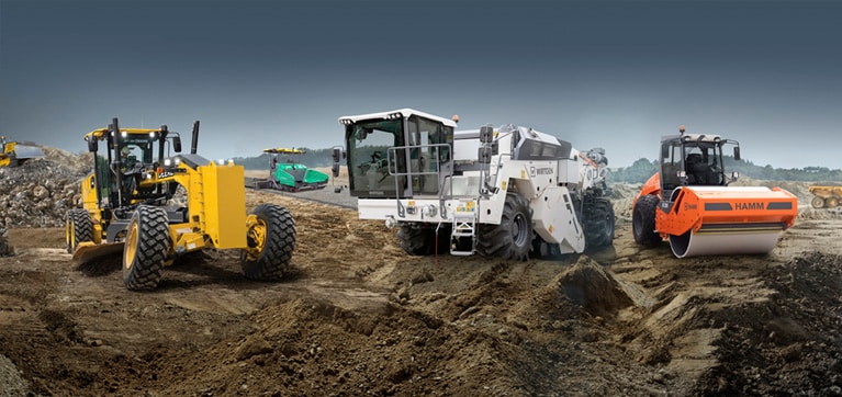 La niveleuse automotrice John Deere aux côtés d'un recycleur à froid Wirtgen et d'un rouleau Hamm avec un finisseur Vögele travaillant en arrière-plan.