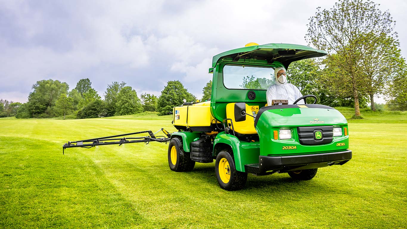 Greenkeeper en tenue de protection traitant la pelouse d’un terrain de golf avec un pulvérisateur John Deere.