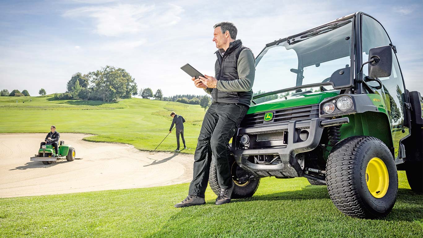 Le greenkeeper en chef vérifie les données sur sa tablette tandis que 2 greenkeepers travaillent sur un terrain de golf en arrière-plan.