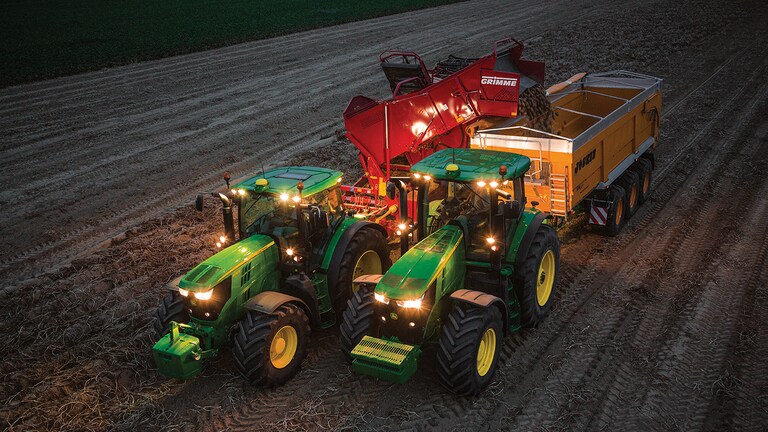 Un tracteur tirant une machine de r&eacute;colte des pommes de terre lu nuit d&eacute;charge des pommes -e terre dans un wagon tir&eacute; par un autre tracteur