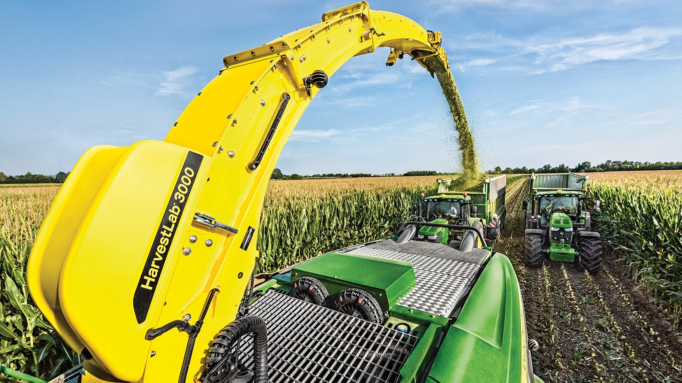 Vue sur l’arrière de l’ensileuse automotrice John Deere série 9000 avec deux wagons d’ensilage tirés par des tracteurs John Deere juste derrière