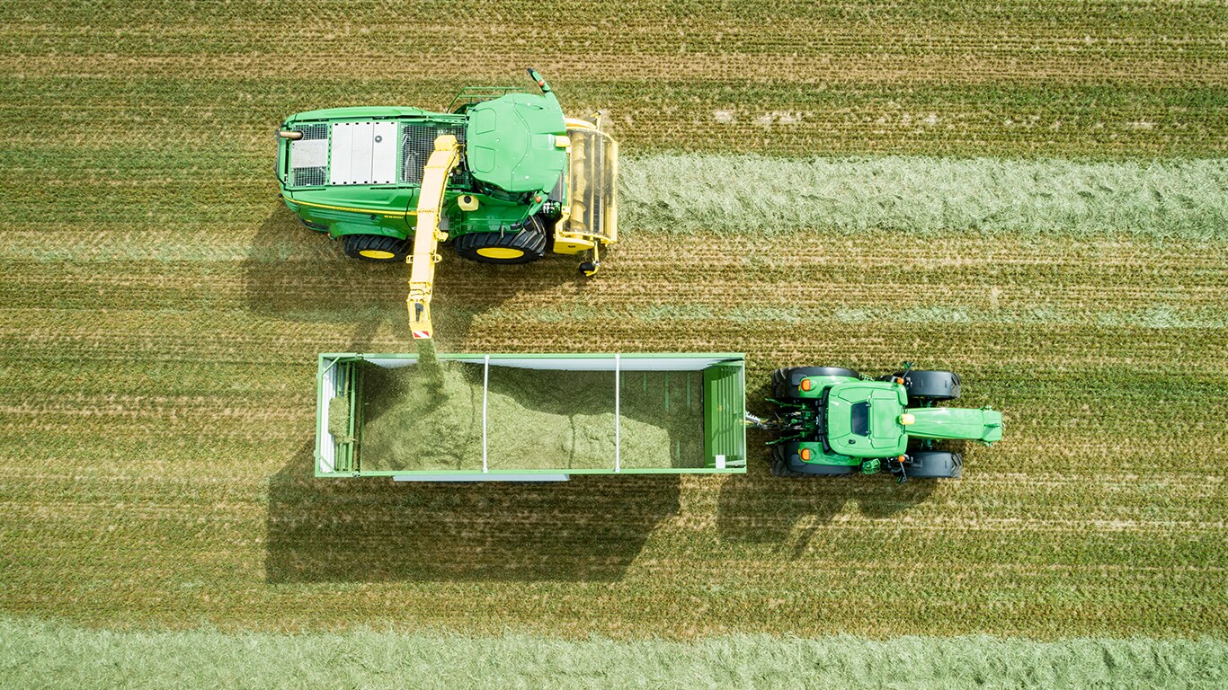 Photo a&eacute;rienne d&rsquo;une ensileuse automotrice John Deere s&eacute;rie 8000 avec un ramasseur &agrave; herbe chargeant de l&rsquo;ensilage dans un wagon tir&eacute; par un tracteur John Deere