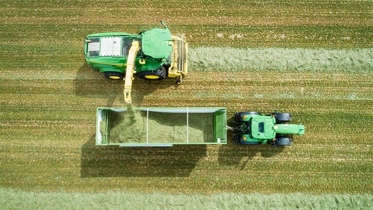 Photo a&eacute;rienne d&rsquo;une ensileuse automotrice John Deere s&eacute;rie 8000 avec un ramasseur &agrave; herbe chargeant de l&rsquo;ensilage dans un wagon tir&eacute; par un tracteur John Deere